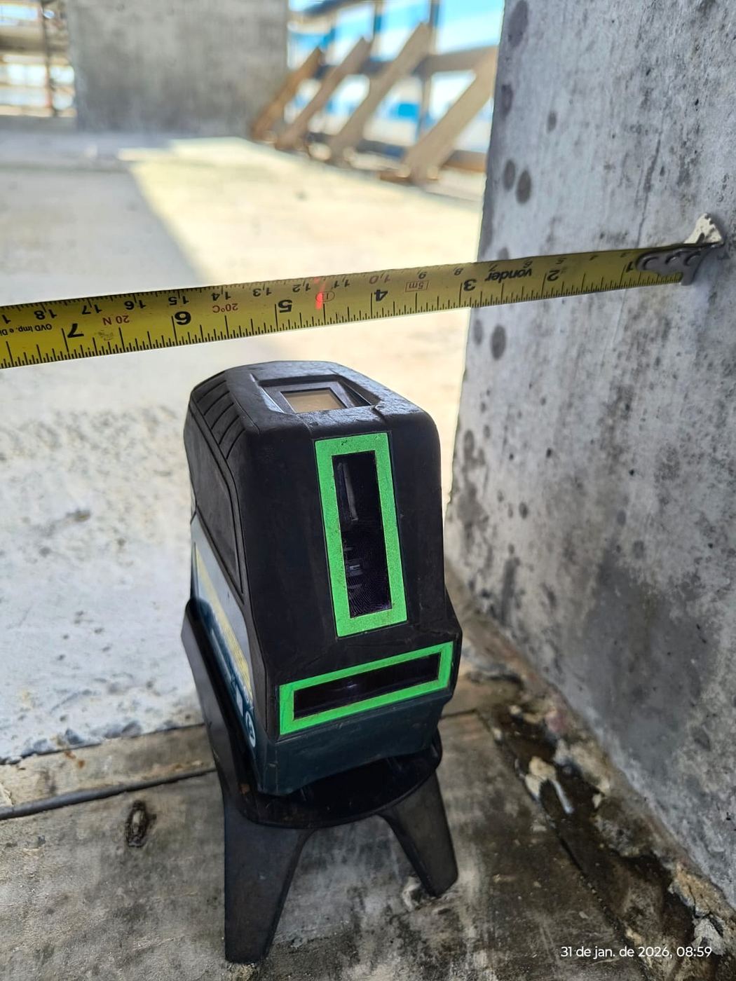 Nível a laser sobre tripé e trena estendida para medição junto a um pilar de concreto.